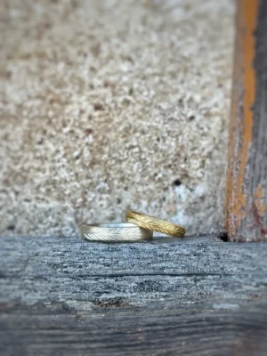 Immergrün Eheringe - fein matte Ringe mit gesägtem Kiefernzweig aus 750 und 585 Gelbgold auf rustikalem Balken