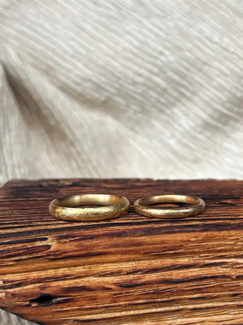 robuste und klassische Sandsturm Eheringe mit grob gefeilter, zerkratzter Struktur aus 750 Gelbgold auf Holz liegend